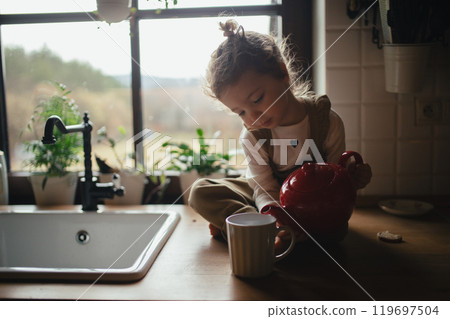 Adorable little girl is pouring tea from the teapot into a cup. The sick girl with cold is sitting on kitchen counter and concentrating. 119697504