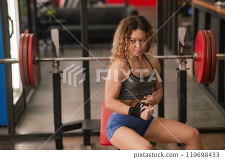 Caucasian forty-year-old woman puts on wrist straps before doing barbell exercises in the gym. Caucasian forty-year-old woman puts on wrist straps before doing barbell exercises in the gym. 119698433