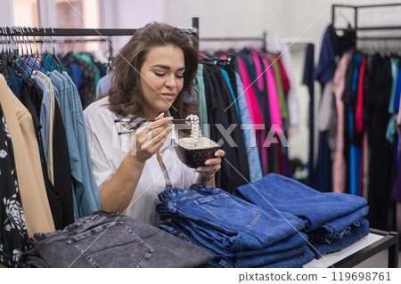 Happy fat woman fooling around and pretending to eat a pearl necklace with Chinese chopsticks in a clothing store for large people. Happy fat woman fooling around and pretending to eat a pearl necklace with Chinese chopsticks in a clothing store for large people. 119698761