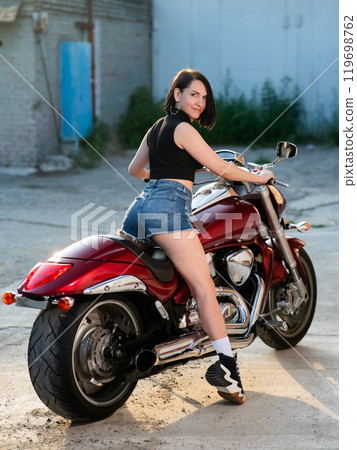 Brunette woman in shorts posing on a red motorcycle. Vertical photo. Brunette woman in shorts posing on a red motorcycle. Vertical photo. 119698762