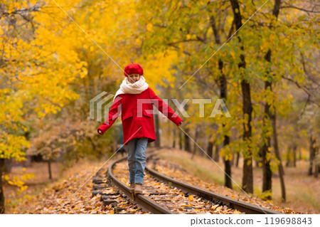 Caucasian girl in a red coat and beret walks along the railway tracks in the park in autumn. Caucasian girl in a red coat and beret walks along the railway tracks in the park in autumn. 119698843