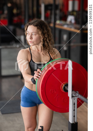 Caucasian forty year old woman putting a weight plate on a barbell in the gym. Vertical photo. Caucasian forty year old woman putting a weight plate on a barbell in the gym. Vertical photo. 119699181