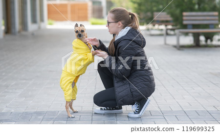 Young Caucasian woman in a jacket walks with a small dog in overalls.  119699325