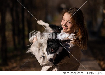 Caucasian woman holding a border collie in her arms while walking in the autumn park. Portrait of a girl with a dog. 119699472