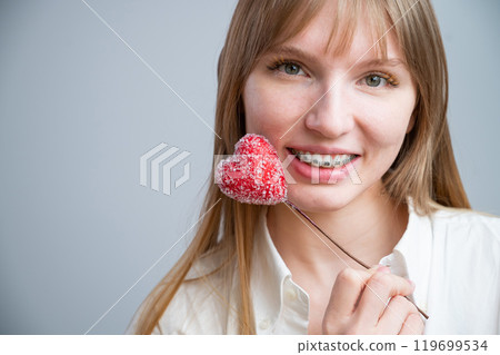 Cute woman with braces on her teeth holds a candy in the form of a heart on white background. Copy space Cute woman with braces on her teeth holds a candy in the form of a heart on white background. Copy space 119699534
