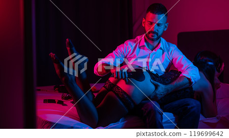 Bearded caucasian man spanks a woman with a leather whip while sitting on a bed in neon light. BDSM sex preferences.  119699642