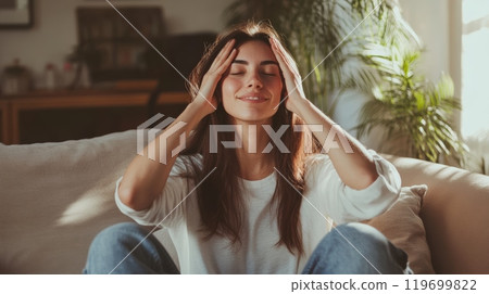 Young woman is sitting on the sofa at home, holding her head in her hands and smiling, enjoying a moment of peace and quiet Young woman is sitting on the sofa at home, holding her head in her hands and smiling, enjoying a moment of peace and quiet 119699822