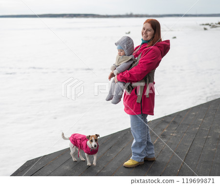 Caucasian woman walks with her son in an ergo backpack and a Jack Russell terrier dog in nature in winter.  119699871