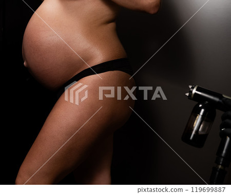 Faceless pregnant woman getting an instant tan. The master applies bronzer with a spray. 119699887