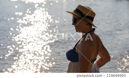 An old woman in a straw hat, sunglasses and a swimsuit is resting on the beach.  119699905