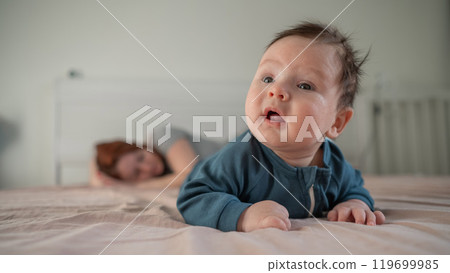 A three-month-old boy lies on his stomach on the bed and his mother sleeps behind him. Postpartum depression. 119699985