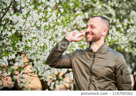 Man allergic using medical nasal drops, suffering from seasonal allergy at spring in blossoming garden. Happy man treating runny nose in front of blooming tree outdoors. Spring allergy concept. 119700562