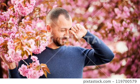 Man allergic suffering from seasonal allergy at spring in blossoming garden at springtime. Bearded man feeling itchy eyes in front of blooming tree. Spring allergy concept. 119700579