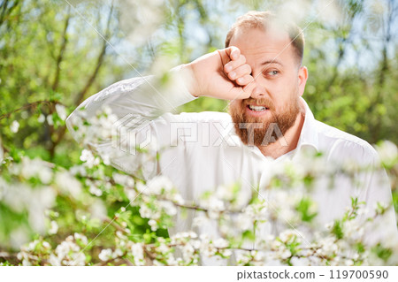 Man allergic suffering from seasonal allergy at spring in blossoming garden at springtime. Close up of bearded man sneezing and feeling itchy eyes in front of blooming tree. Spring allergy concept. Man allergic suffering from seasonal allergy at spring in blossoming garden at springtime. Close up of bearded man sneezing and feeling itchy eyes in front of blooming tree. Spring allergy concept. 119700590