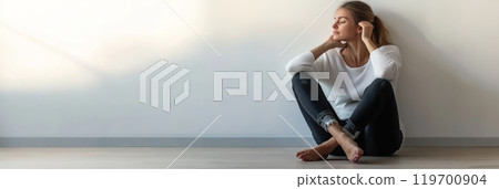 Young woman is sitting on the floor with her back against the wall, eyes closed, enjoying a moment of peace and quiet 119700904