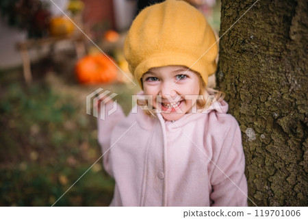 A cheerful child in a yellow hat smiles while peeking around a tree in the autumn woods 119701006