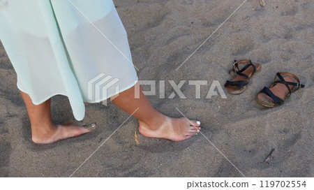barefoot girl in blue dress walking on gray fine sand and abandoned black leather sandals nearby, barefoot walk on beach, girl's legs on sandy shore barefoot girl in blue dress walking on gray fine sand and abandoned black leather sandals nearby, barefoot walk on beach, girl's legs on sandy shore 119702554