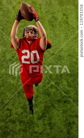 Top view image of concentrated man, American football player in red uniform in motion on stadium, catching ball with precision 119703358