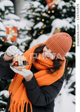 Smiling woman holding festive Christmas burger with ornaments and wrapped gift. Holiday food creativity, winter celebrations, and outdoor festive fashion Smiling woman holding festive Christmas burger with ornaments and wrapped gift. Holiday food creativity, winter celebrations, and outdoor festive fashion 119703676