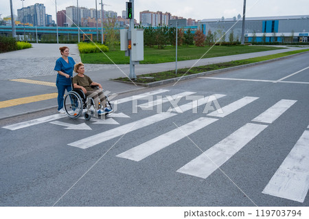 Red-haired nurse pushing an elderly woman in a wheelchair across the road. Red-haired nurse pushing an elderly woman in a wheelchair across the road. 119703794