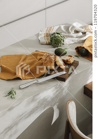 Sliced bread on wooden board, avocado, artichoke, linen towel and stainless still kitchen knife. Kitchen aesthetic concept Sliced bread on wooden board, avocado, artichoke, linen towel and stainless still kitchen knife. Kitchen aesthetic concept 119704320