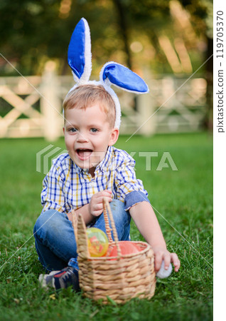 A cheerful young boy wearing bunny ears is sitting on the grass with a wicker basket filled with colorful Easter eggs. A cheerful young boy wearing bunny ears is sitting on the grass with a wicker basket filled with colorful Easter eggs. 119705370