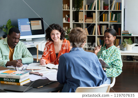 Group of diverse colleagues working together on project around table, sharing ideas and engaging in discussion. Books and computer on table, representing teamwork and productivity 119707635