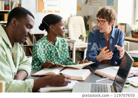 Coworkers discussing while collaborating on a work project. Open books and notebooks on table indicate an ongoing study or research conversation Coworkers discussing while collaborating on a work project. Open books and notebooks on table indicate an ongoing study or research conversation 119707650