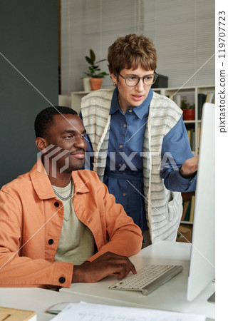 Caucasian woman and African American man collaborating on project in modern office, both expressing engagement and teamwork while standing and sitting at desk with computer 119707723