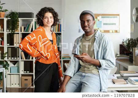 Smiling colleagues collaborating in office setting with shelves and plants in background creating a modern and relaxed atmosphere with gadgets and paperwork on the desk 119707828