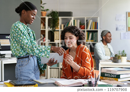 Two women collaborating on academic project, one showing tablet to other. Various books and academic supplies visible, with another person studying in background 119707908