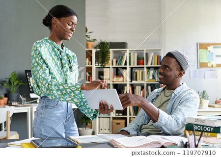 Two people collaborating and discussing project details using a tablet in modern office environment surrounded by books and work tools 119707910