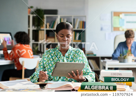 Portrait of African American woman focus on her work in modern office space with books on desk and colleagues in background engaging in various activities Portrait of African American woman focus on her work in modern office space with books on desk and colleagues in background engaging in various activities 119707924