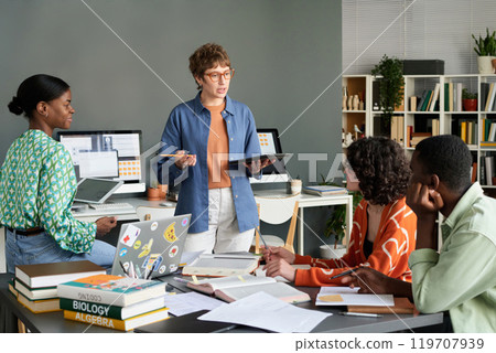 Multiracial team collaborating at office, using laptops and tablets while engaging in lively discussion. Books and documents on the table, suggesting a productive work environment 119707939