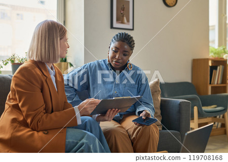 Portrait of adult African American woman talking to female finance expert explaining tax forms and showing clipboard with documents Portrait of adult African American woman talking to female finance expert explaining tax forms and showing clipboard with documents 119708168