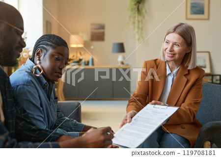 Portrait of smiling mature woman handing clipboard with contract or legal forms to African American couple in home interior copy space Portrait of smiling mature woman handing clipboard with contract or legal forms to African American couple in home interior copy space 119708181