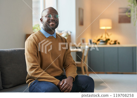Portrait of adult African American man wearing glasses and smiling at camera while sitting on couch in minimal home setting copy space 119708258