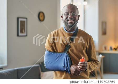 Waist up portrait of smiling adult African American man with broken arm in sling looking at camera and holding bottle of pills copy space Waist up portrait of smiling adult African American man with broken arm in sling looking at camera and holding bottle of pills copy space 119708269