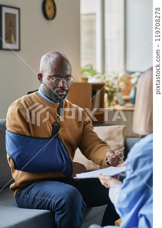 Vertical portrait of adult African American man with broken arm signing medical form on clipboard during at home doctors visit Vertical portrait of adult African American man with broken arm signing medical form on clipboard during at home doctors visit 119708278