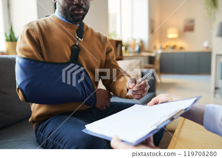 Close up of African American man with arm in sling signing medical form on clipboard during at home emergency doctors visit copy space Close up of African American man with arm in sling signing medical form on clipboard during at home emergency doctors visit copy space 119708280