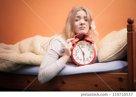 Unhappy girl in bed with alarm clock Unhappy girl in bed with alarm clock 119710537