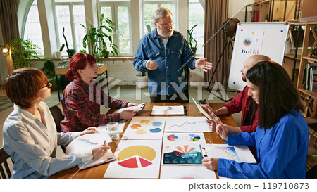 Elderly team leader uses visual data, charts, and graphs on whiteboard to explain business strategies during collaborative team meeting 119710873