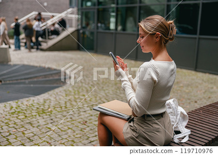 A Stylish Young Woman Engaged in Texting While Enjoying the Outdoors in a City Setting A Stylish Young Woman Engaged in Texting While Enjoying the Outdoors in a City Setting 119710976