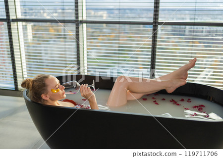Spa relaxation. Woman lying in bath with holding glass with red wine. Girl relaxing in bathroom at home. Pretty female taking hot bath drinking wine. Stress relief. Rest after hard working day 119711706