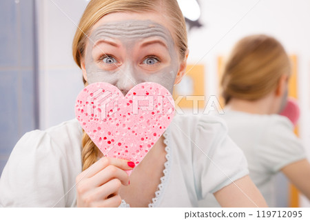 Woman having grey face mask holding heart sponge Woman having grey face mask holding heart sponge 119712095