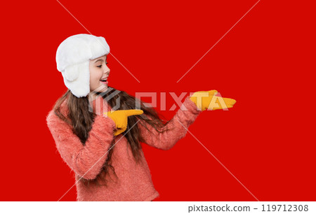 Girl wearing trendy outfit with knitted beanie hat. Girl with winter hat isolated on red. Winter fashion style trend. Teen girl in hat enjoying childhood. Point finger 119712308