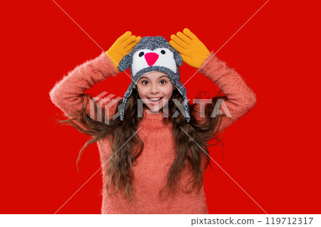Teen kid girl wearing warm knitted winter earflap hat and sweater isolated on red Teen kid girl wearing warm knitted winter earflap hat and sweater isolated on red 119712317