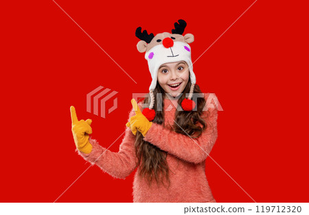 Teenage girl in Xmas hat isolated on red. Winter fashion for teenage style. Girl in knitted hat in winter season. Childhood lifestyle. Winter child wardrobe. Point finger up Teenage girl in Xmas hat isolated on red. Winter fashion for teenage style. Girl in knitted hat in winter season. Childhood lifestyle. Winter child wardrobe. Point finger up 119712320