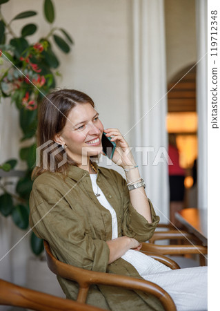 A cheerful woman talks on the phone in a cozy setting, surrounded by stylish decor and plants 119712348