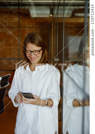 A Smiling Woman Using a Smartphone in a Modern Office, Radiating Positivity and Professionalism A Smiling Woman Using a Smartphone in a Modern Office, Radiating Positivity and Professionalism 119712414